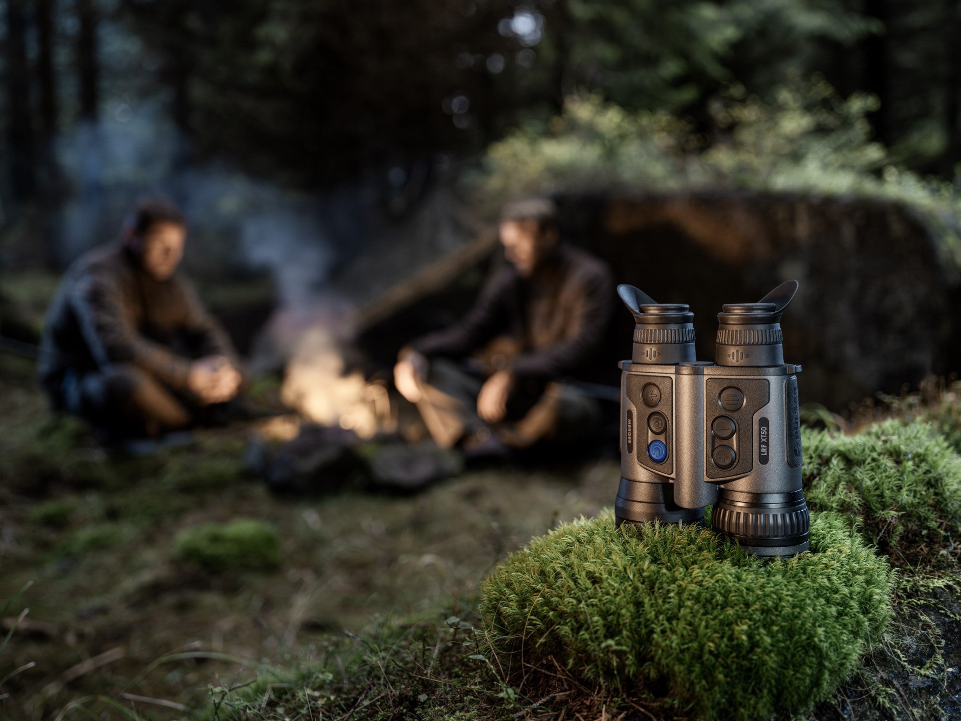 Binoculars on a mossy rock with people around a campfire in the background.